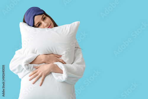 Young woman in pajamas and sleeping mask holding pillow on blue background photo