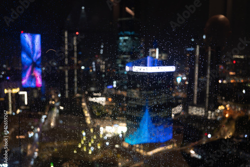 Photography Nighttime view of Kuala Lumpur through a rain-covered window, featuring blurred city lights and vibrant colors, creating a dynamic and textured atmosphere