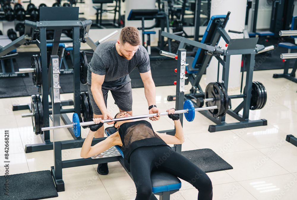 A man is helping a woman lift weights. The man is wearing a black shirt and gray shorts. A man is helping a woman lift weights. The man is wearing a black shirt and gray shorts.