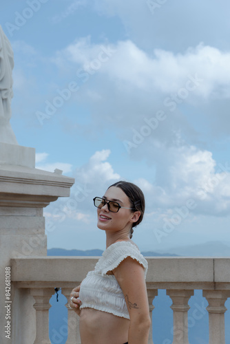 A woman in a white crop top and black shorts smiles while standing next to a classical statue on a stone balcony. The blue sky with scattered clouds complements her cheerful expression