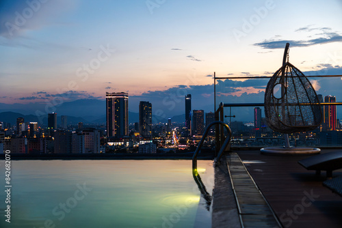 Sunset view of Da Nang, Vietnam, with high-rise buildings and a rooftop pool. A hanging chair adds a cozy element to the serene cityscape as lights begin to illuminate the skyline.