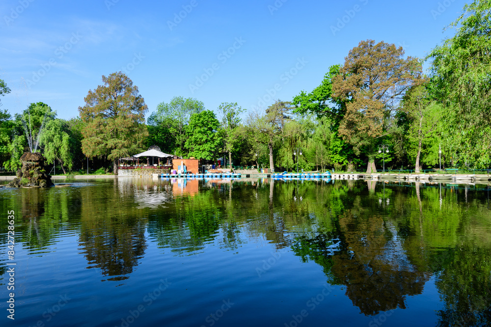Vivid green landscape with old large linden trees and small boats near the lake in Cismigiu Garden (Gradina Cismigiu), a public park in the city center of Bucharest, Romania, in a sunny spring day. Vivid green landscape with old large linden trees and small boats near the lake in Cismigiu Garden (Gradina Cismigiu), a public park in the city center of Bucharest, Romania, in a sunny spring day.