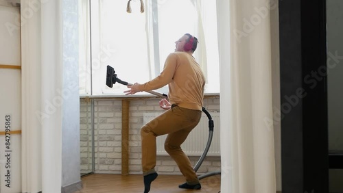 A handsome man dances with a vacuum cleaner. House cleaning with dancing. A young man cleans his apartment while listening to music on his headphones