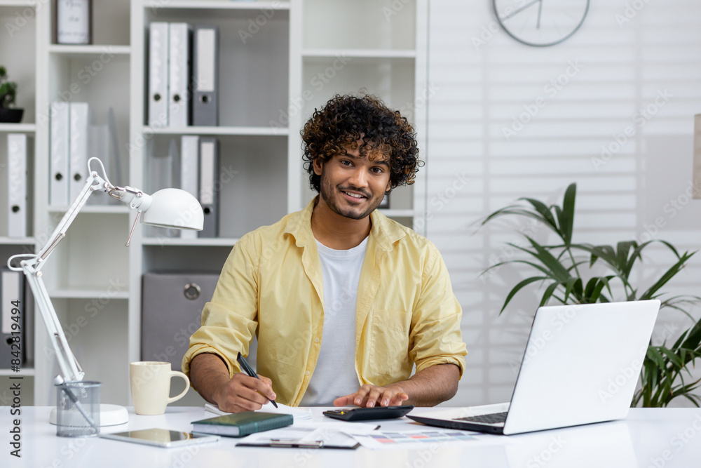 Young professional working at a desk in a modern home office, using a laptop and organizing paperwork with a smile Young professional working at a desk in a modern home office, using a laptop and organizing paperwork with a smile