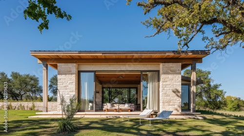 Wallpaper Mural Small rectangular two-story country house with light-colored concrete and wood beams, large front windows, covered patio seating, in Texas' rustic landscape with green grass and trees under clear blue Torontodigital.ca