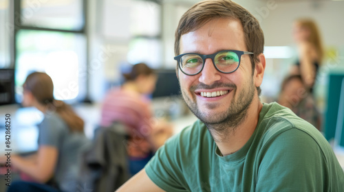 A cheerful young man with glasses and a green t-shirt smiles confidently in a brightly lit office, colleagues working in the blurred background.