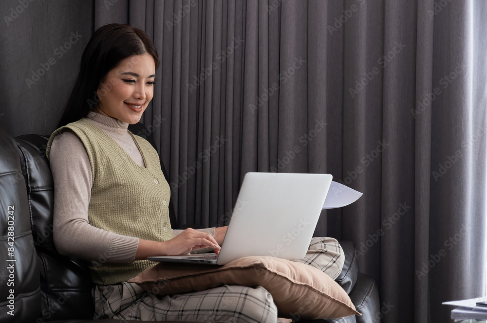 Smiling Asian woman using laptop on sofa, Concept of comfortable remote work Smiling Asian woman using laptop on sofa, Concept of comfortable remote work