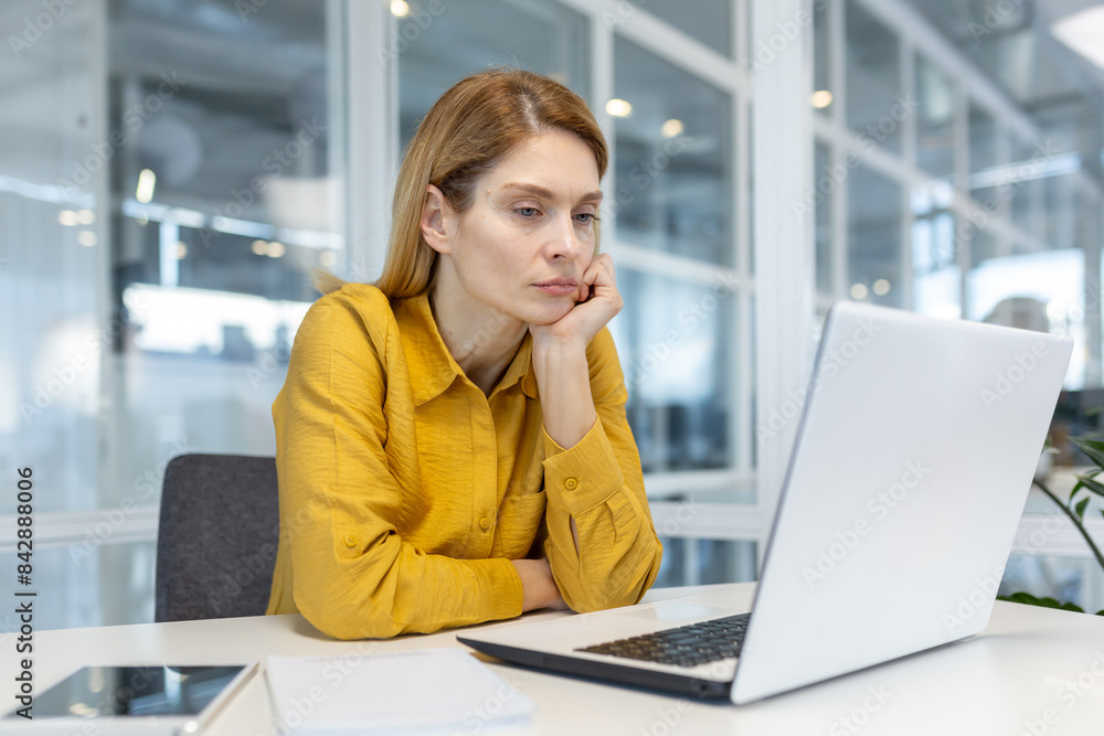 Woman in a yellow shirt, focused and engaged, working on a laptop in a bright, modern office setting. Woman in a yellow shirt, focused and engaged, working on a laptop in a bright, modern office setting.