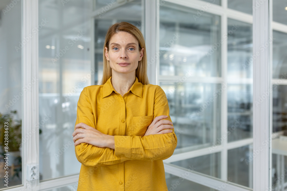 Confident businesswoman wearing a yellow shirt stands with arms crossed in a modern office setting, looking directly at the camera Confident businesswoman wearing a yellow shirt stands with arms crossed in a modern office setting, looking directly at the camera