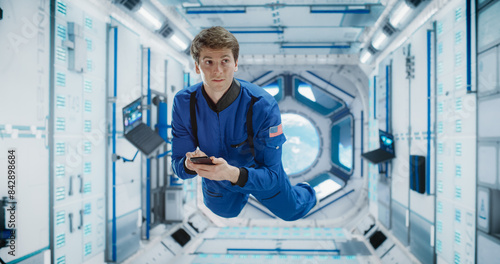 Cheerful Male Astronaut Floating in Zero Gravity Inside a Spaceship. Happy Caucasian Specialist Using Smartphone in Space, Checking News, and Communicating with Family and Friends Online.