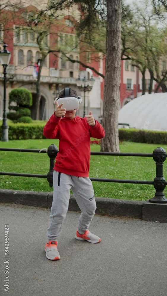 A young boy in a red hoodie puts on a virtual reality helmet and starts playing video games in a park, modern technology A young boy in a red hoodie puts on a virtual reality helmet and starts playing video games in a park, modern technology