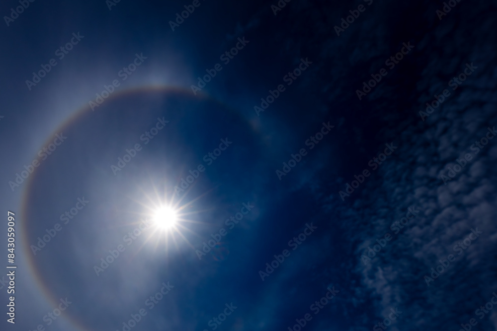 Obraz premium A captivating image of a sun halo against a deep blue sky, created by the refraction of sunlight through ice crystals in the atmosphere. The sun is bright, surrounded by a circular rainbow-like halo.