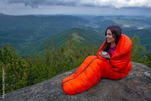 Woman tourist in orange sleeping bag with coffee cup on the rock in Carpathian mountains photo