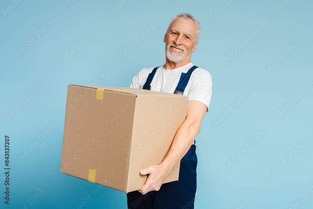 Cheerful senior courier carrying heavy cupboard box at blue wall background Cheerful senior courier carrying heavy cupboard box at blue wall background