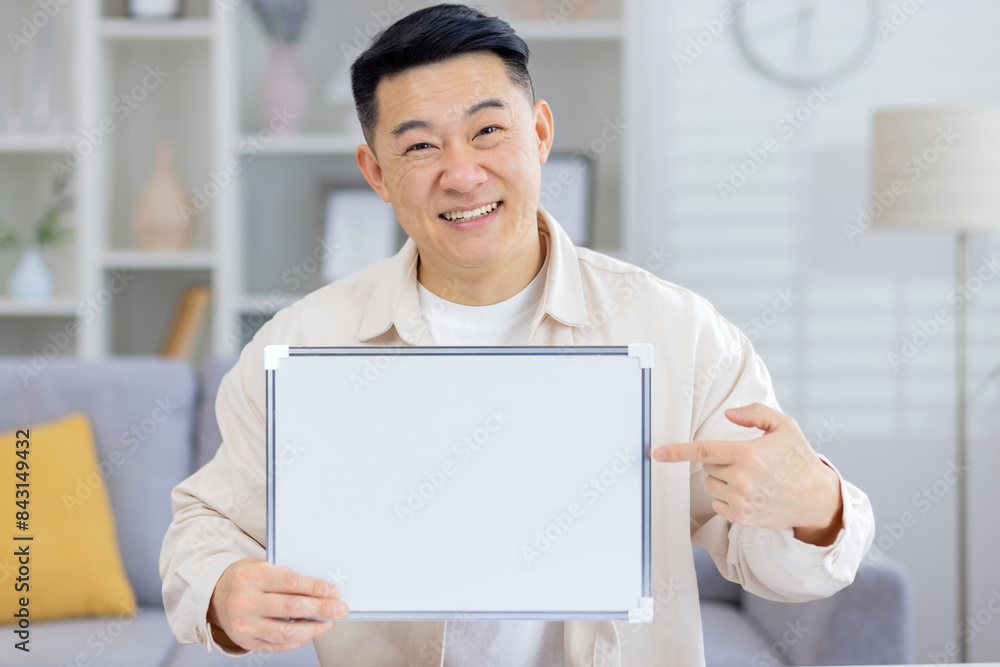 Smiling man holding a blank sign while pointing and looking cheerful in a cozy living room. Perfect for messages and advertising concepts. Smiling man holding a blank sign while pointing and looking cheerful in a cozy living room. Perfect for messages and advertising concepts.