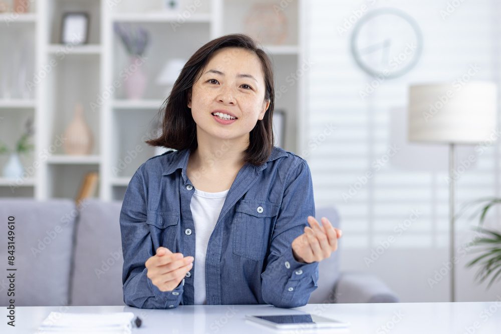Asian woman engaging in a video call, chatting and communicating from a modern living room setting. Asian woman engaging in a video call, chatting and communicating from a modern living room setting.