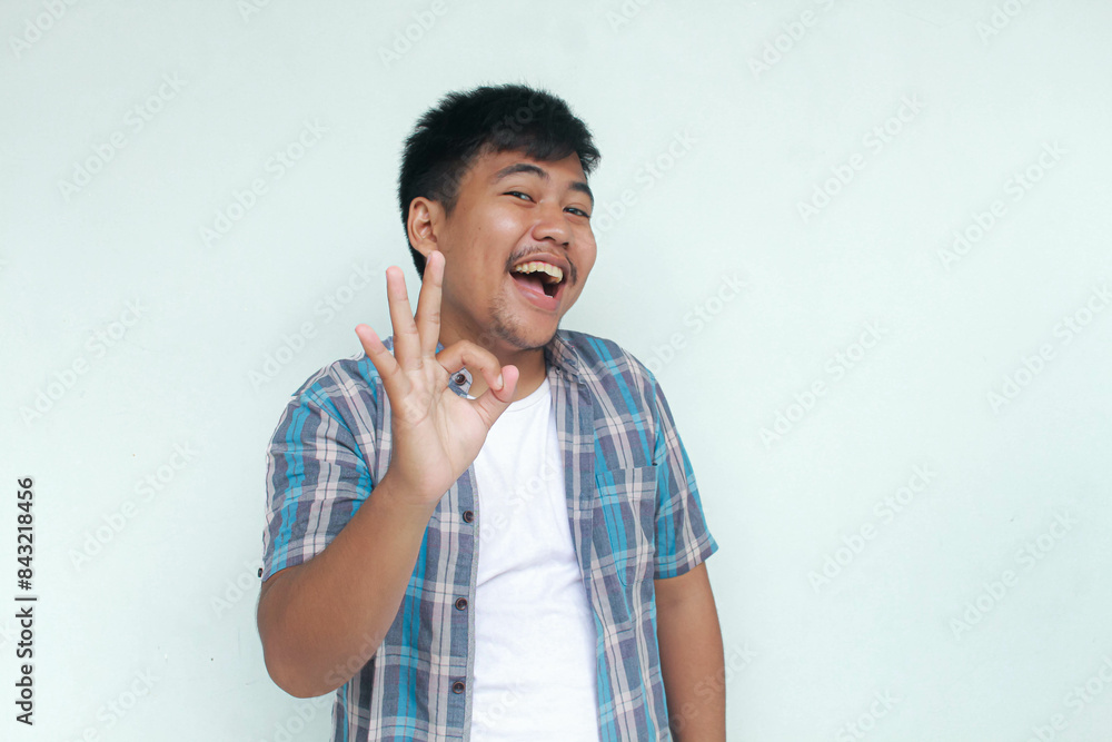 Excited and happy young Asian man gives thumbs up or ok sign hand gesture of approval, isolated by gray background. Excited and happy young Asian man gives thumbs up or ok sign hand gesture of approval, isolated by gray background.