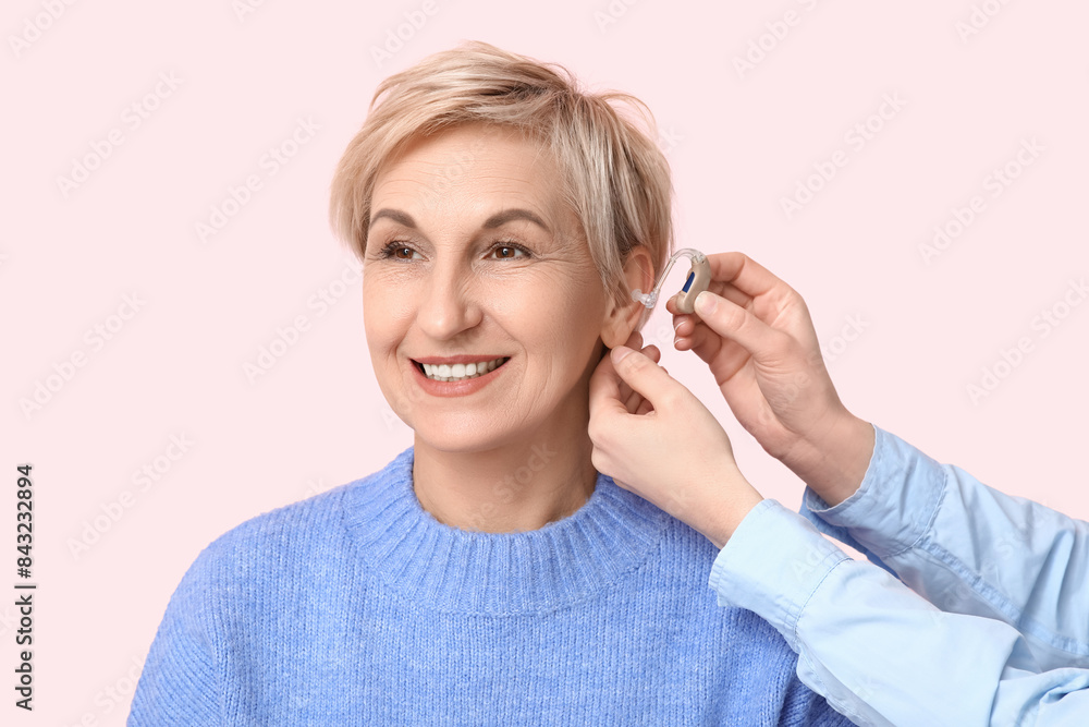 Young woman putting hearing aid in mature woman's ear on pink background Young woman putting hearing aid in mature woman's ear on pink background