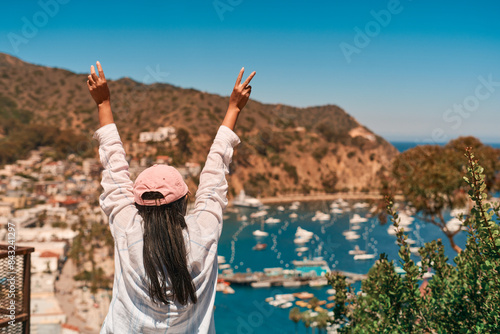 Back view of a happy Asian girl in a pink hat with arms raised and a peace sign standing on a hill of Catalina Island, CA, in summer and viewing the ocean and boats at Avalon Harbor  from above