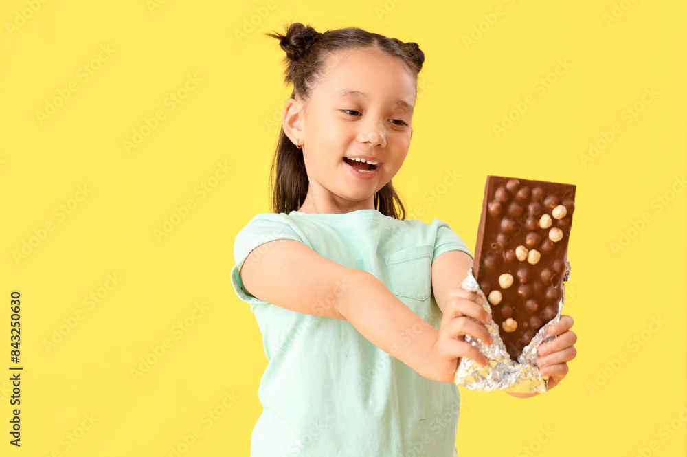 Cute little Asian girl with chocolate bar on yellow background