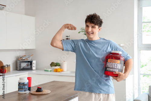 Sporty young man with protein powder showing muscles in kitchen