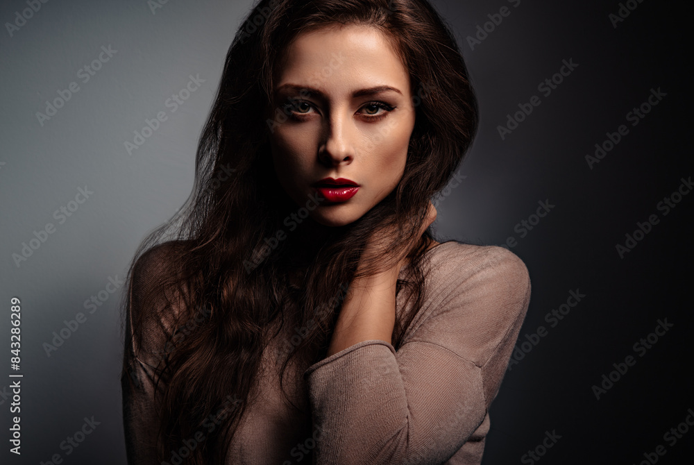 Sexy makeup with red lipstick woman holding long brown curly hair and posing in elegant dress with vamp look on studio background. Half shadow and light. Closeup Sexy makeup with red lipstick woman holding long brown curly hair and posing in elegant dress with vamp look on studio background. Half shadow and light. Closeup