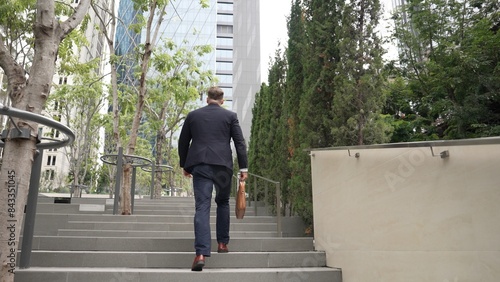 Back view of caucasian manager with formal suit walking up stairs while holding his suitcase in green city. Professional business man going to workiplace while climb stair in eco urban city. Urbane.. photo