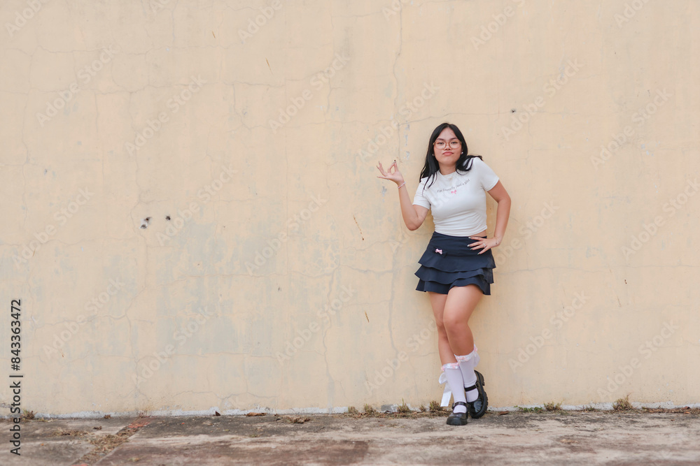 Young asian woman in schoolgirl inspired attire stands confidently against an aged wall outdoors, making an ok sign. Young asian woman in schoolgirl inspired attire stands confidently against an aged wall outdoors, making an ok sign.