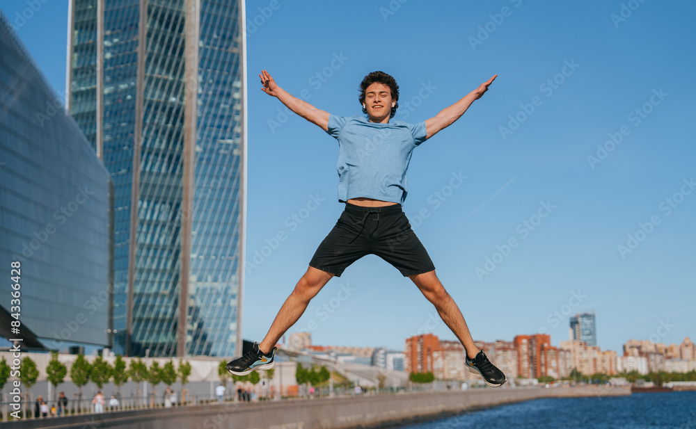 Man jumping with arms and legs outstretched, wearing a blue t-shirt and black shorts. He is outdoors by the waterfront with a modern cityscape and clear blue sky in the background Man jumping with arms and legs outstretched, wearing a blue t-shirt and black shorts. He is outdoors by the waterfront with a modern cityscape and clear blue sky in the background