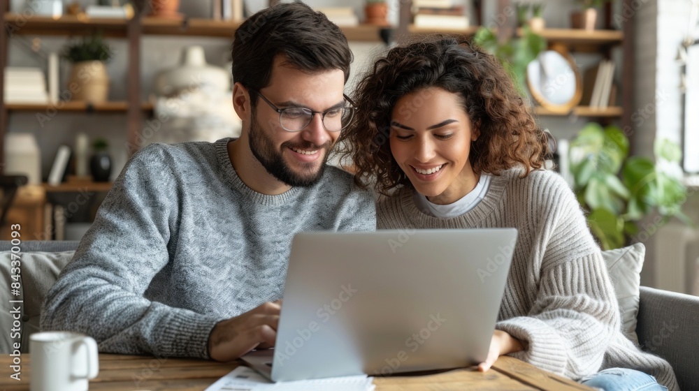 A couple sits together on a couch, sharing a laptop and looking at something on the screen A couple sits together on a couch, sharing a laptop and looking at something on the screen