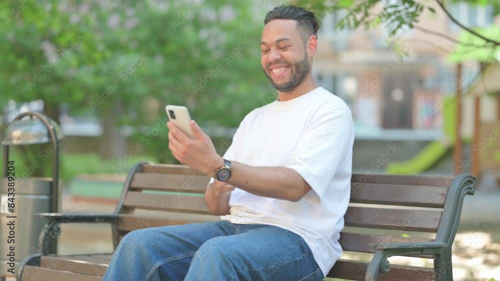 Young Man Celebrating on Phone while Sitting Outdoor on Bench Young Man Celebrating on Phone while Sitting Outdoor on Bench