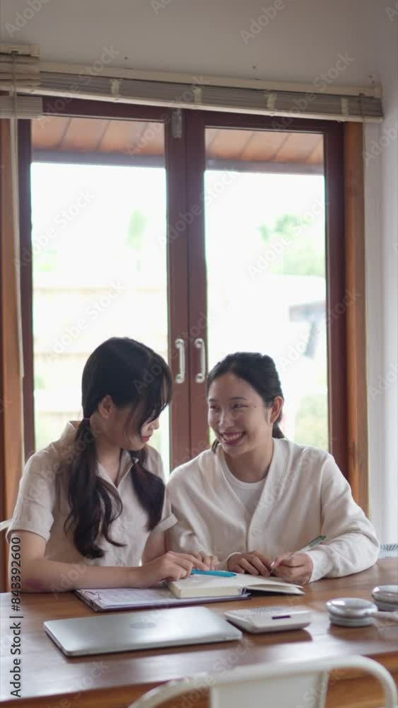 Two Asian university female friends are studying together by discussing the course content and explaining it through books and study materials to help their friend understand at her home. Two Asian university female friends are studying together by discussing the course content and explaining it through books and study materials to help their friend understand at her home.