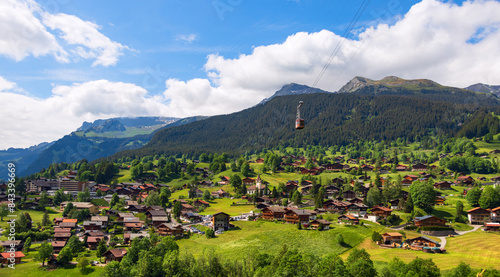 Breathtaking panoramic view of the alpine village of Grindelwald in a valley surrounded by bright green mountain slopes and the old cable car with a cabin on the mountain slopes background 