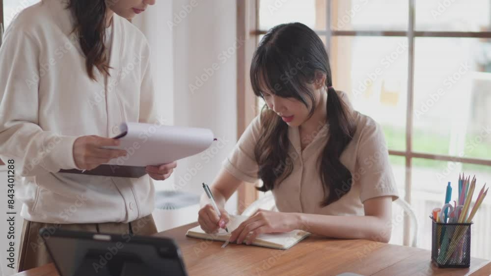 Two Asian university female friends are studying together by discussing the course content and explaining it through books and study materials to help their friend understand at her homeใ Two Asian university female friends are studying together by discussing the course content and explaining it through books and study materials to help their friend understand at her homeใ