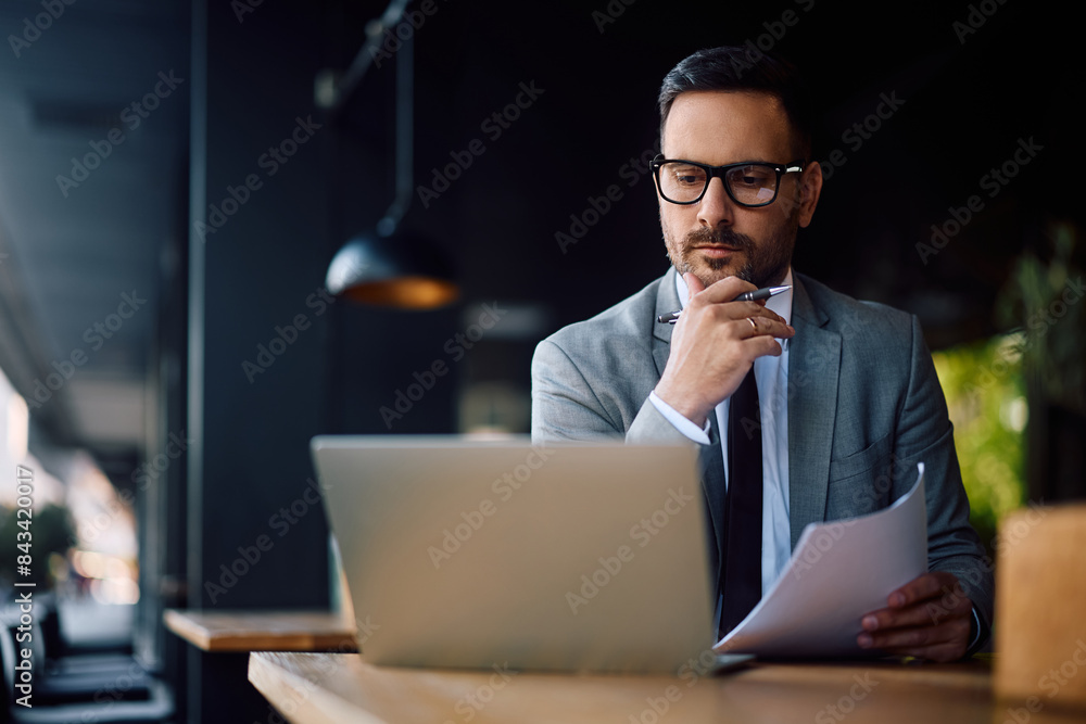 Pensive entrepreneur using laptop while analyzing business reports in café. Pensive entrepreneur using laptop while analyzing business reports in café.