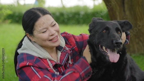 Portrait of happy beautiful Asian woman petting and cuddling cute east european black female shepherd, showing care, love and friendship while relaxing with dog in nature.