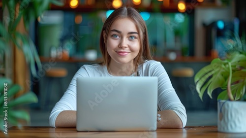 Young woman with a pleasant smile working on her laptop, possibly in a cafe or co-working space, surrounded by indoor plants.