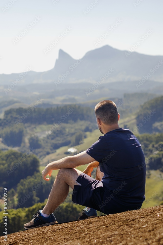 Naklejka premium A young man sits with his back turned on a rock at the peak of a mountain, a valley stretches below, and a striking rock formation is visible on the horizon.