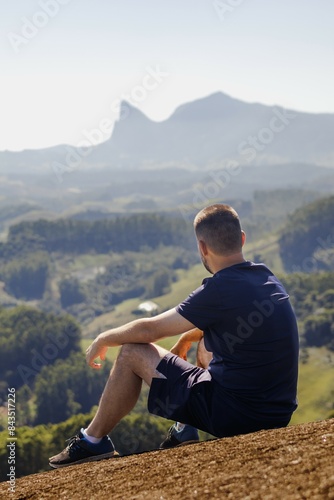 A young man sits with his back turned on a rock at the peak of a mountain, a valley stretches below, and a striking rock formation is visible on the horizon. © Raquele