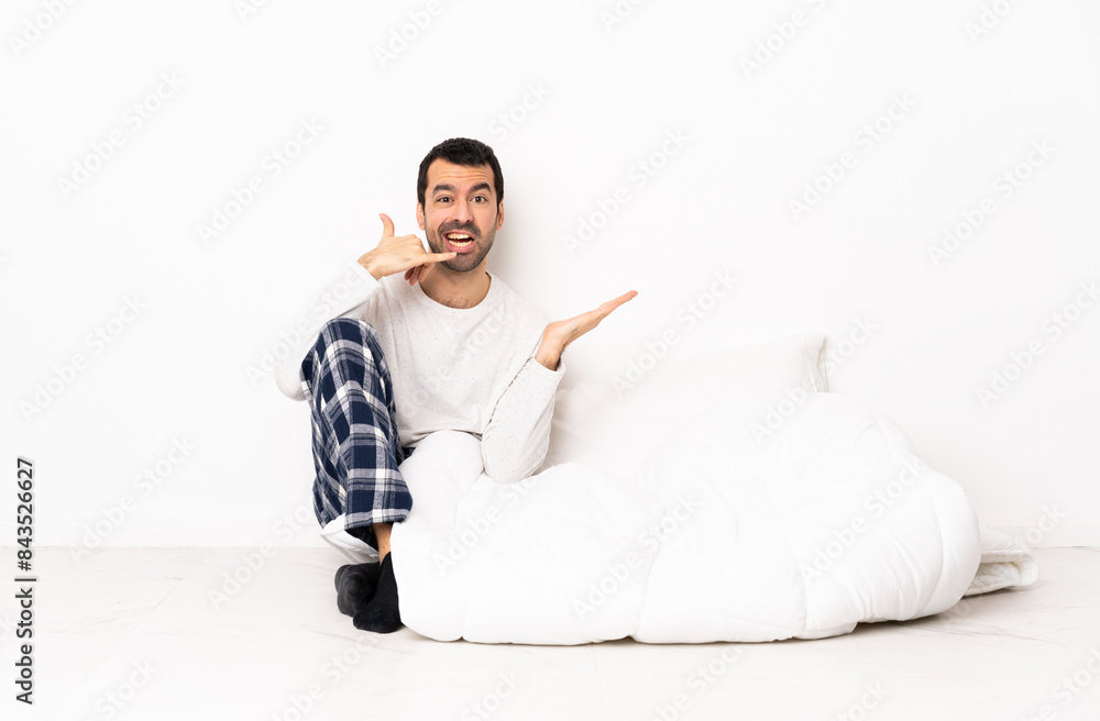 Caucasian man in pajamas sitting on the floor at indoors making phone gesture and doubting Caucasian man in pajamas sitting on the floor at indoors making phone gesture and doubting