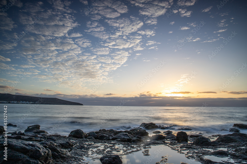 View of the sunrise over the Atlantic Ocean. Fantastic clouds and a bay with a stone beach on a volcanic island. Cold lava stones by the sea at Playa de Tarajalejo beach, Fuerteventura, Canary Islands View of the sunrise over the Atlantic Ocean. Fantastic clouds and a bay with a stone beach on a volcanic island. Cold lava stones by the sea at Playa de Tarajalejo beach, Fuerteventura, Canary Islands