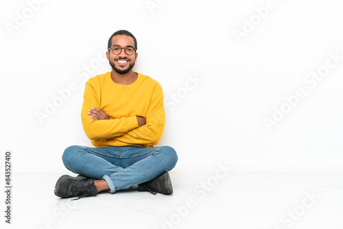 Young Ecuadorian man sitting on the floor isolated on white wall keeping the arms crossed in frontal position