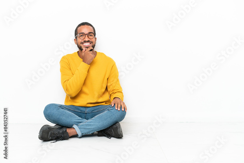 Young Ecuadorian man sitting on the floor isolated on white wall with glasses and smiling
