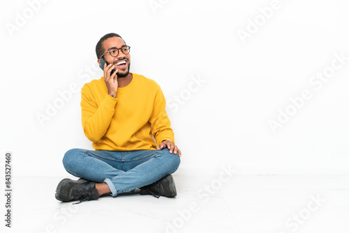 Young Ecuadorian man sitting on the floor isolated on white wall keeping a conversation with the mobile phone