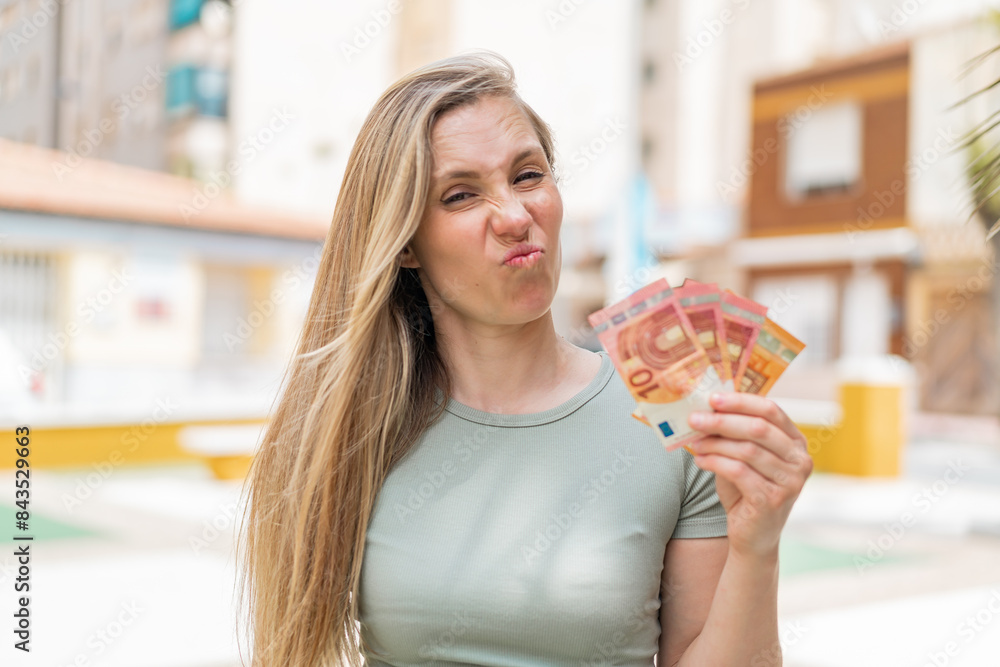 Young blonde woman taking a lot of money at outdoors with sad expression Young blonde woman taking a lot of money at outdoors with sad expression