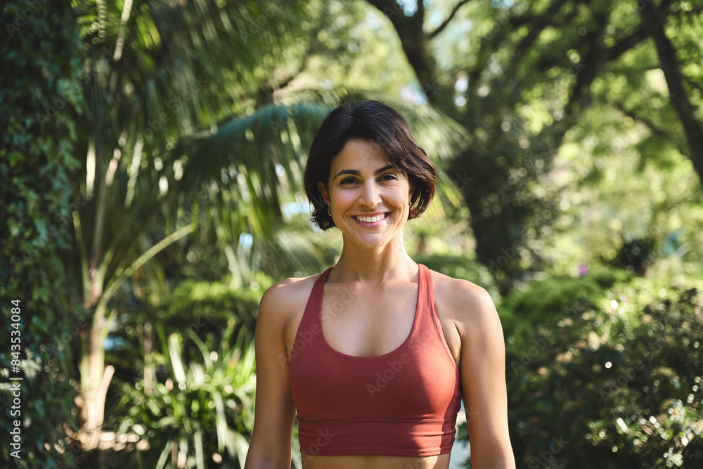 Happy smiling fit sporty active slim confident young Hispanic woman wearing sportswear top enjoying summer sunny day looking at camera standing in green nature park or forest. Portrait. Happy smiling fit sporty active slim confident young Hispanic woman wearing sportswear top enjoying summer sunny day looking at camera standing in green nature park or forest. Portrait.