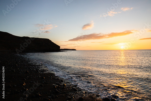 View of the sunrise over the Atlantic Ocean. View over a stone beach of a volcanic island. Cold lava stones by the sea on the beach at Cueva de Playa en Tarajalejo, Fuerteventura, Canary Islands, Spai