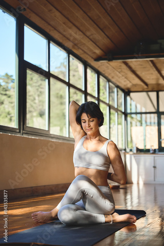 Young fit sporty slim healthy Hispanic yogi woman wearing grey top leggings doing yoga stretching training exercises sitting in yoga pose on mat in modern light yoga gym studio. Vertical candid photo.