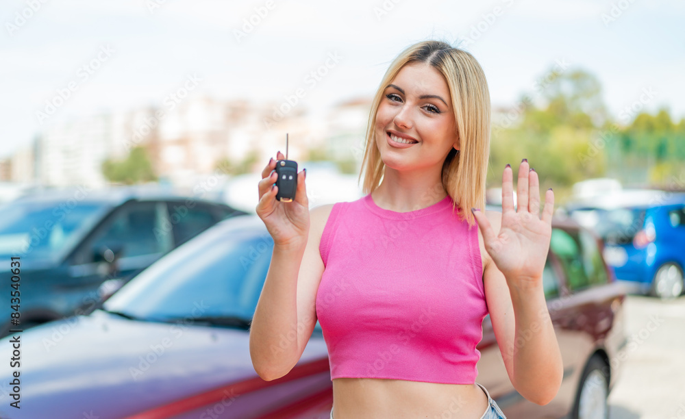 Young pretty blonde woman holding car keys at outdoors saluting with hand with happy expression Young pretty blonde woman holding car keys at outdoors saluting with hand with happy expression