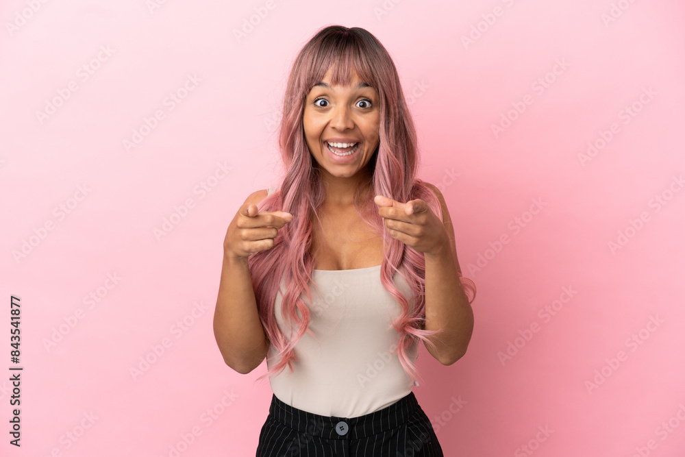 Young mixed race woman with pink hair isolated on pink background surprised and pointing front Young mixed race woman with pink hair isolated on pink background surprised and pointing front